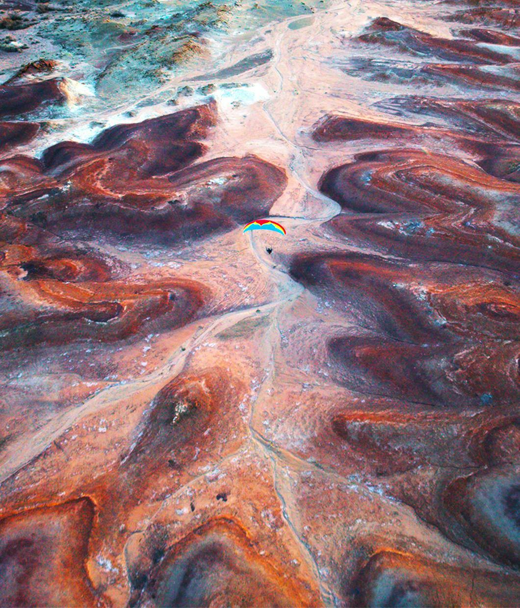 Ein Paragleiter fliegt über einer Steinwüste. Die Farben der Steine sind rötlich und braun. Der Paragleiter ist hebt sich durch seine Farben, Blau, Rot und Gelb von der Steinwüste ab. © ©BANFF Moving Adventures
